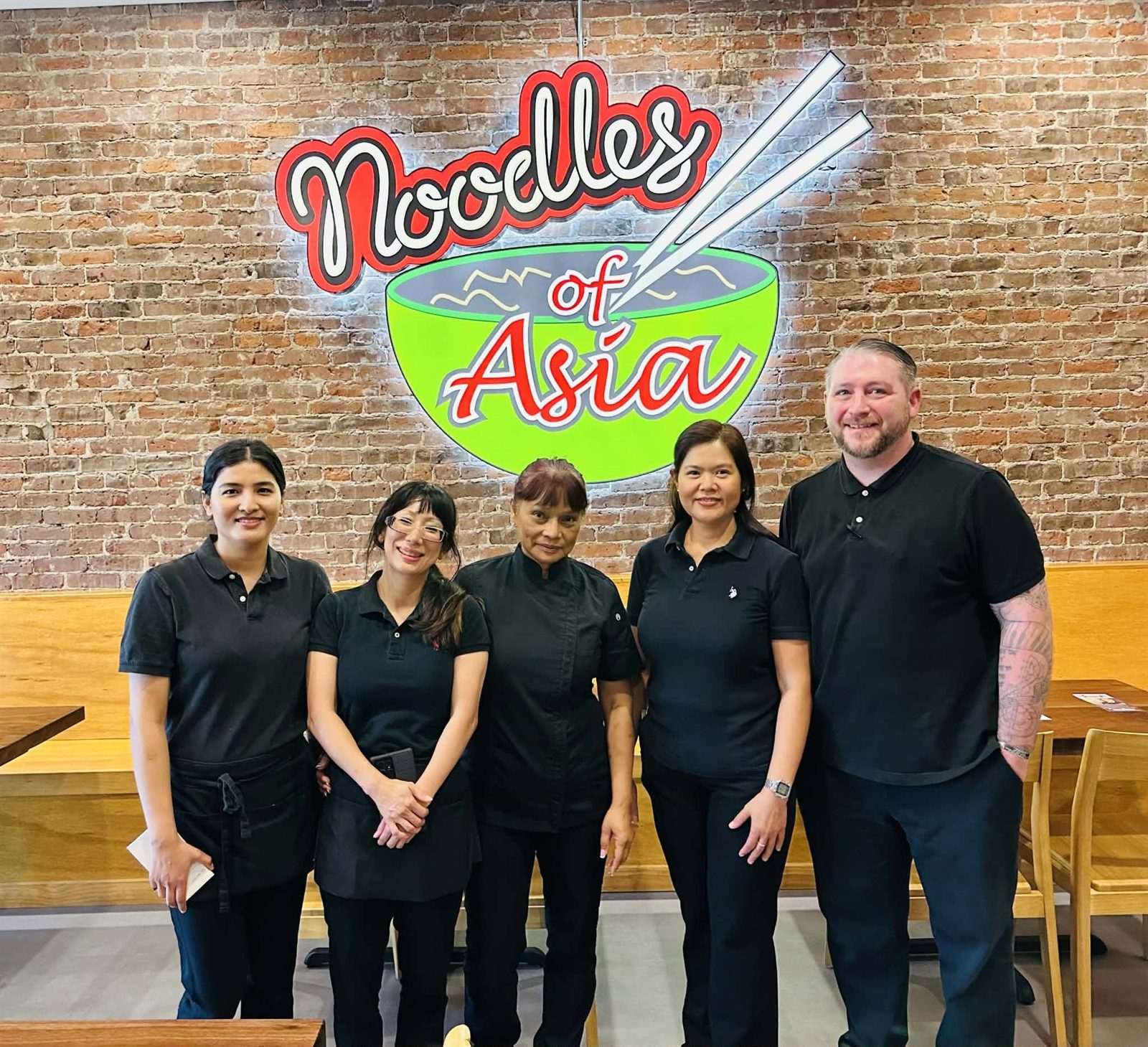 The Noodles of Asia team standing beneath the lit sign on the brick wall